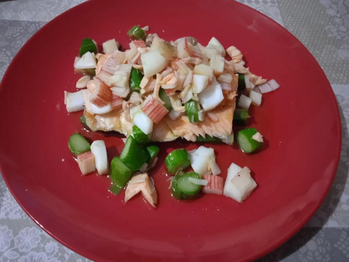 Salmón hecho a la plancha al que le hemos cubierto con un poco de salpicón de pescado y marisco que hemos piuesto en un plato llano de color rojo listo para servir.