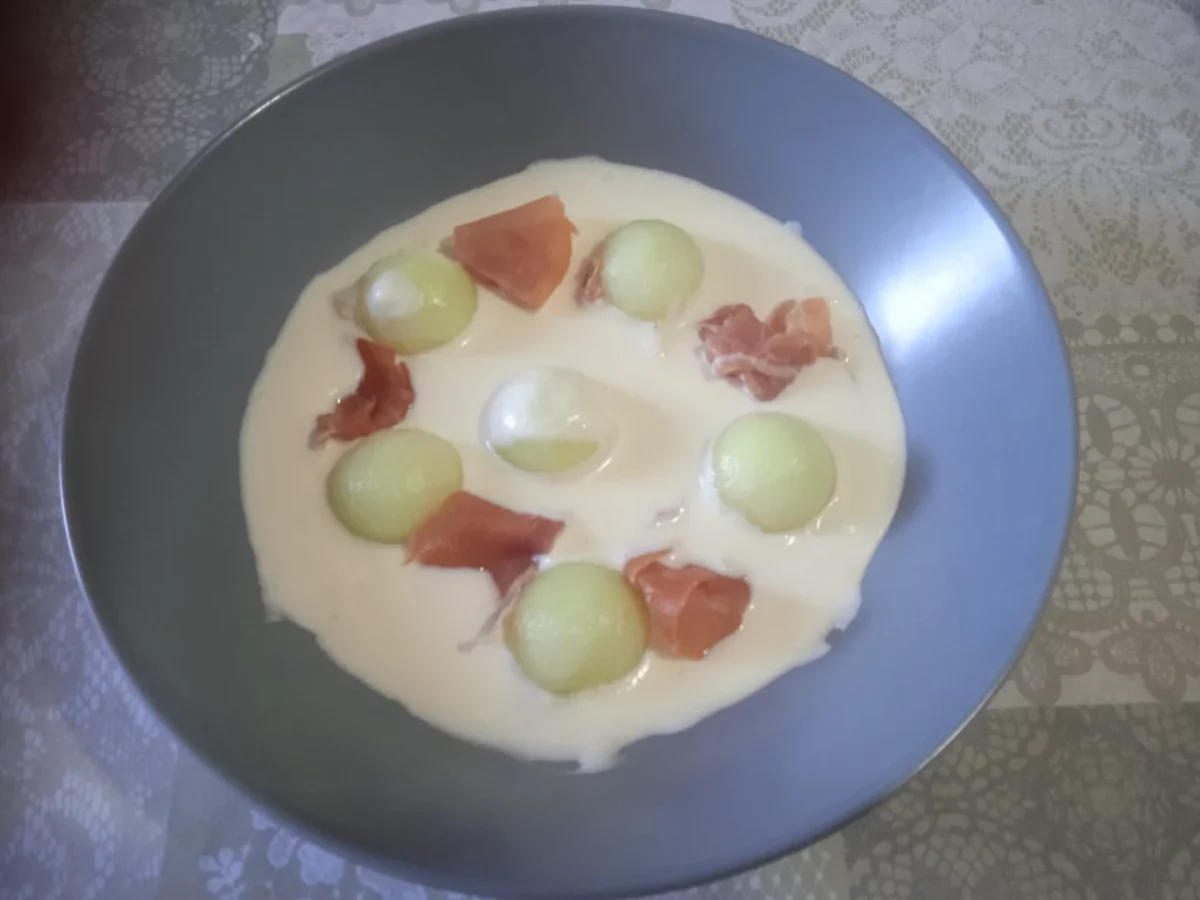 receta de crema de melón puesta en un plato hondo de color azúl con unas bolas de melón y trozos de jamón serrano lesta para servirla.