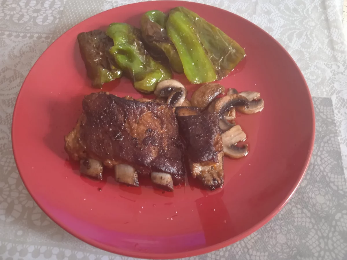 Costillas de cerdo marinadas acabadas puestas en un plato llano de color rojo con una guarnición al lado de pimientos fritos.
