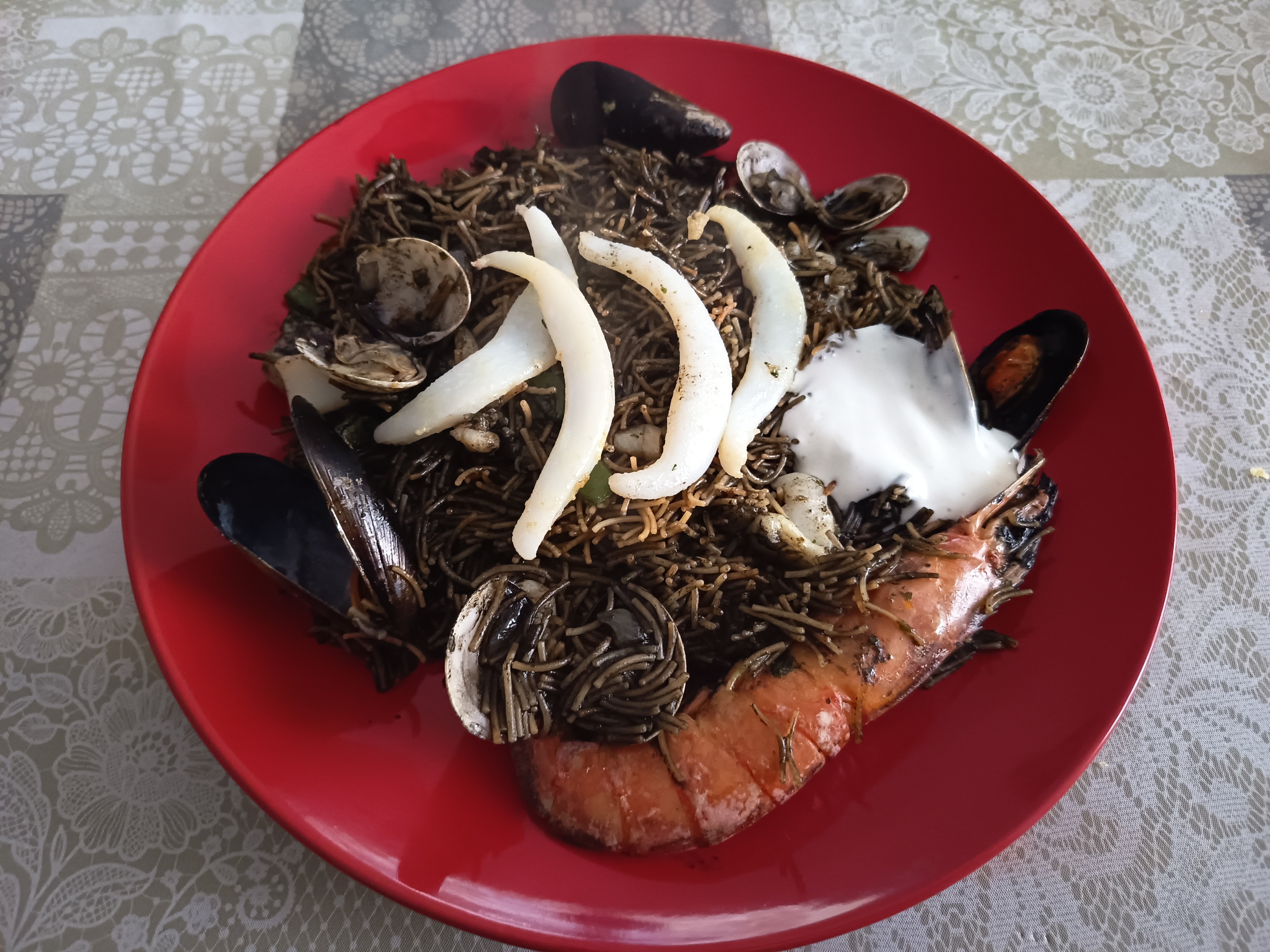 Receta de fideuá negra acabada colocada en un plato de color rojo con una gamba, almejas, mejillones y tiras de sepia cortadas finas y hechas a la plancha