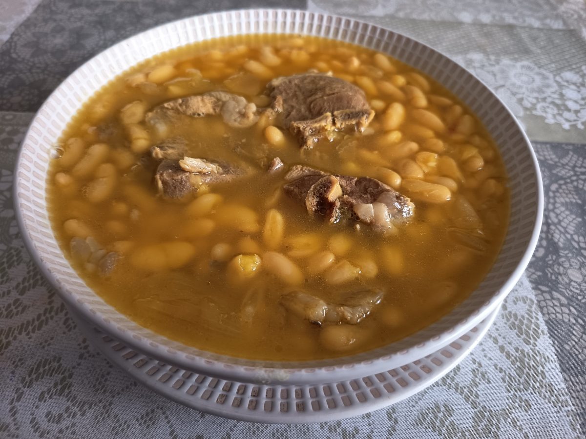 Receta de Judías con cordero acabada colocada en un plato hondo con 4 trozosde cordero puesta en un hule de color beis con motivos florales.