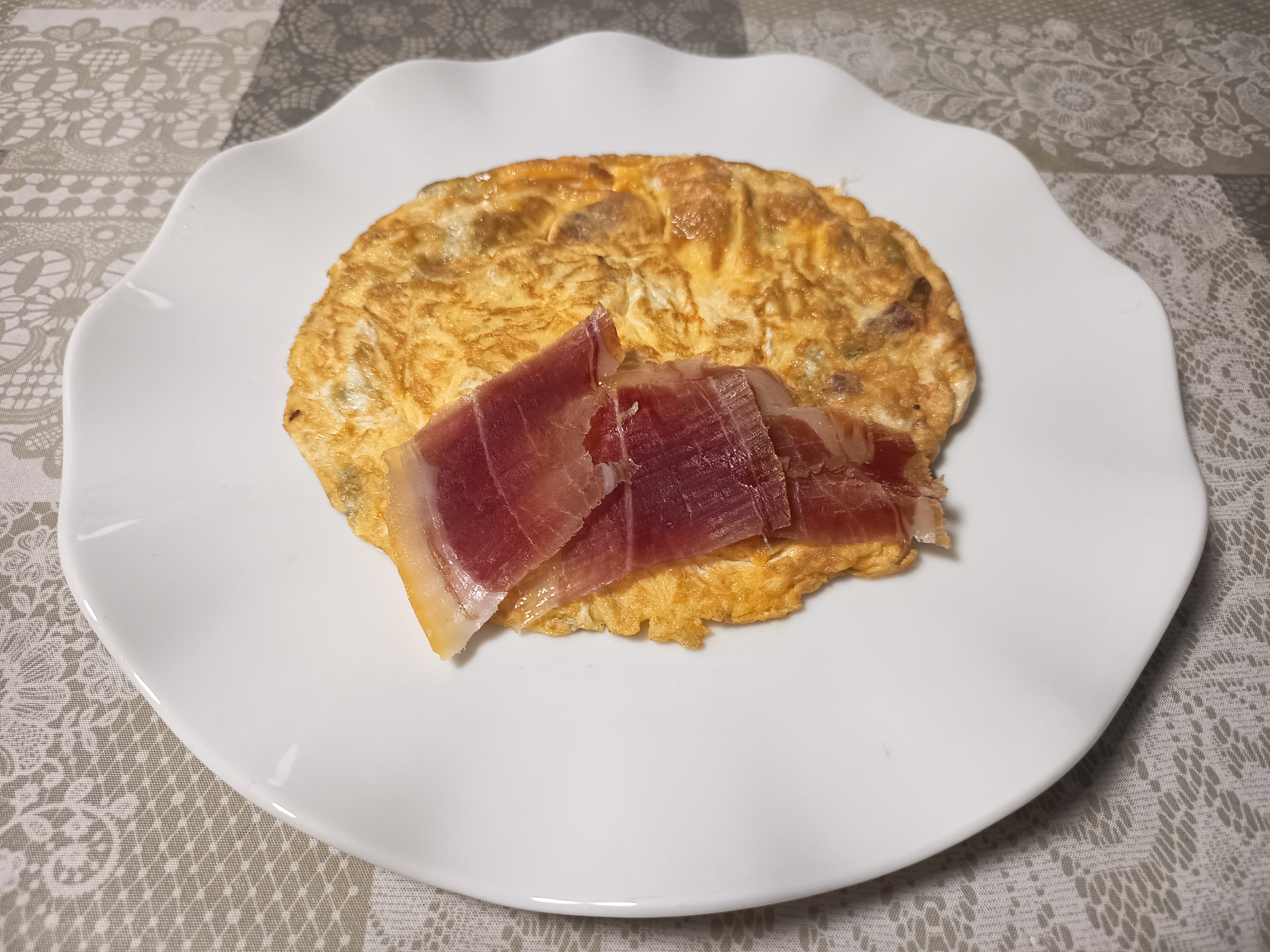 Tortilla de ajos tiernos y gambas acabada y puesta en un plato llano de color blanco con dos trozos de jamón serrano sobre la tortilla puesta en un hule de dolor gris con motivos florales.