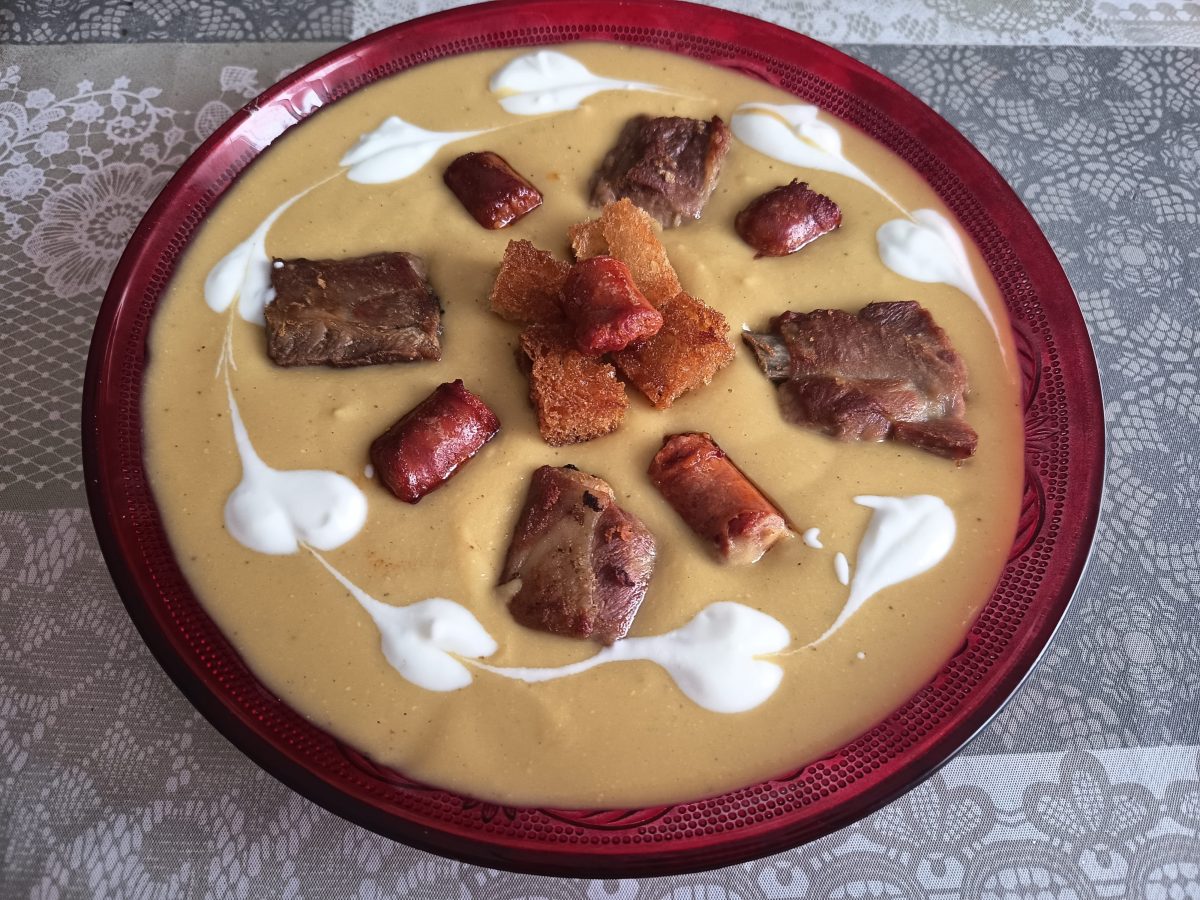 Crema de lentejas, puestas en un plato llano con unos trozos de costilla de cerdo y chorizo y en el centro unos costrones de pan frito; alrededor, unos corazones de nata líquida, puesto en un mantel de color gris con ornamentos florales.