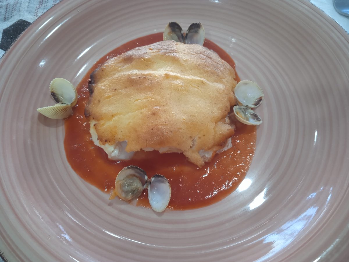 recetade bacalao a la muselina dealioli acabada puestasobre una base de salsa de tomate y alrededor unos berberchos abiertos en un plato llano de color morado claro.