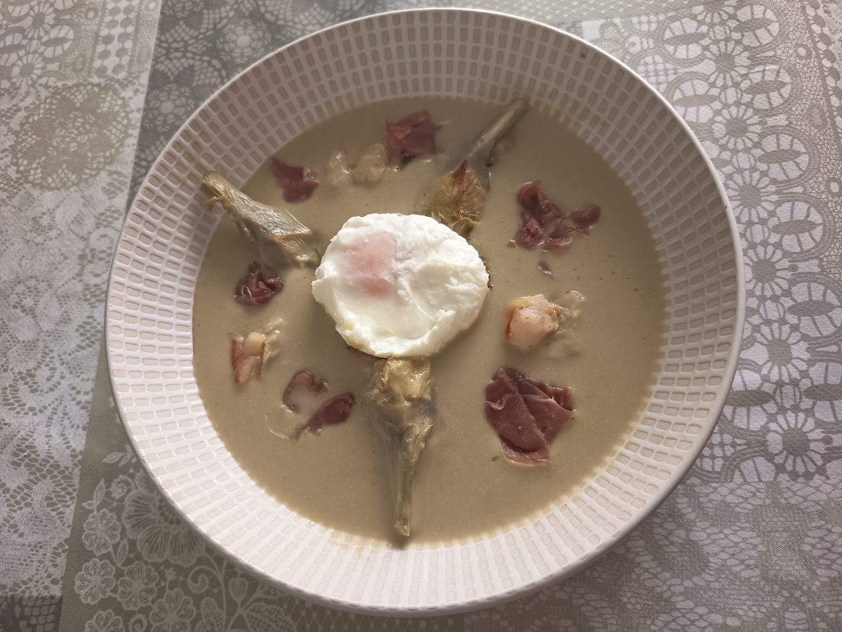 Receta de la crema de alcachofas con huevo poche, jamón serrano y gambas puesto en un plato hondo lista para servirla.